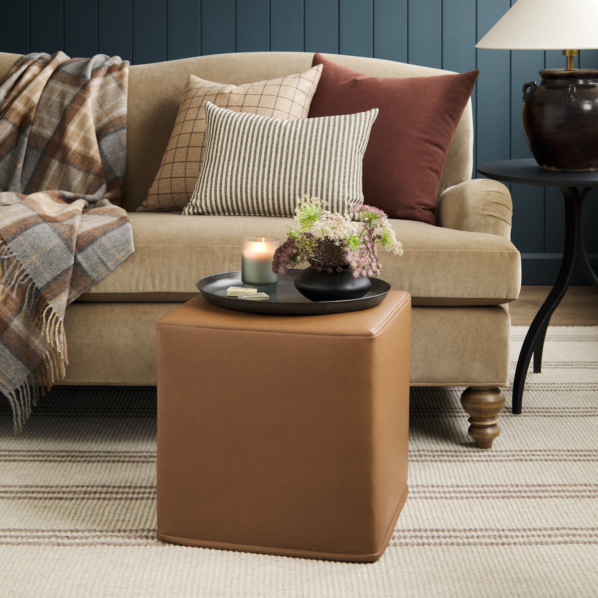 Tonic Living Smythe Saddle faux leather cube ottoman styled as an accent table in a warm living room with a neutral sofa and layered pillows.